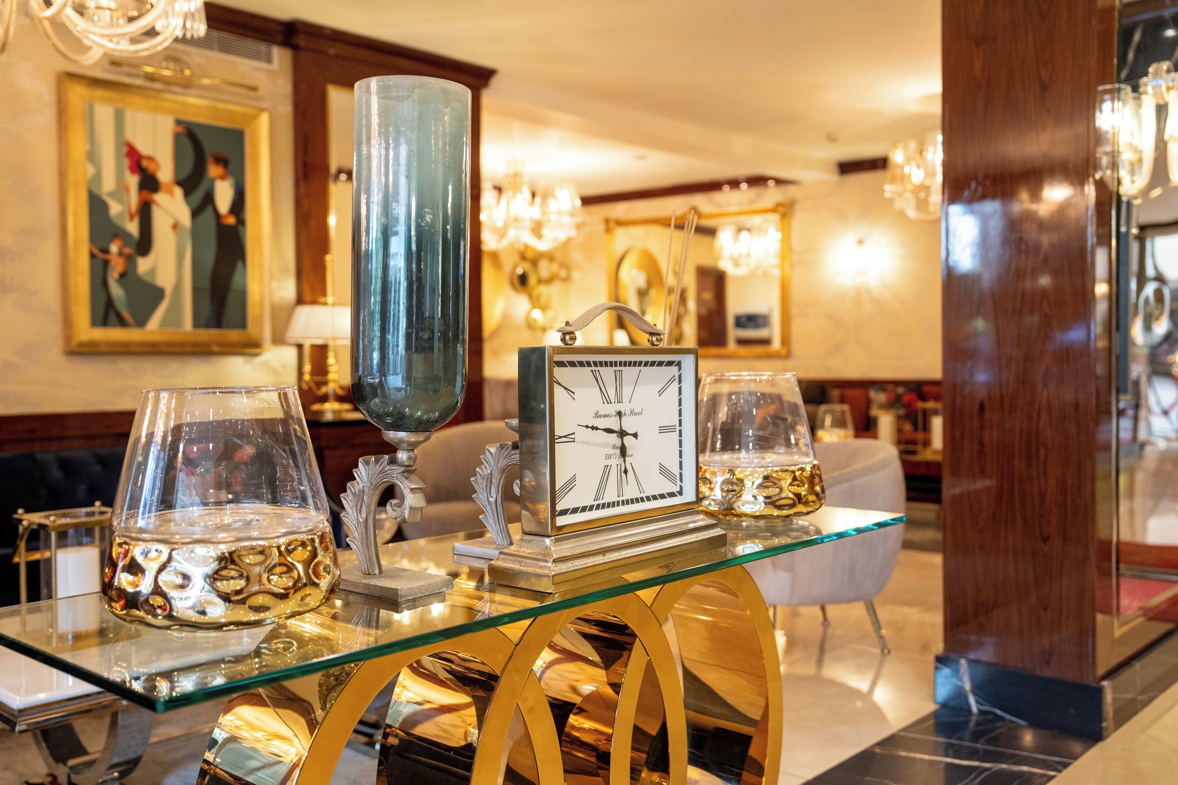 A glass-top table with a clock, decorative glasses, and a vase in a well-lit room with a chandelier.