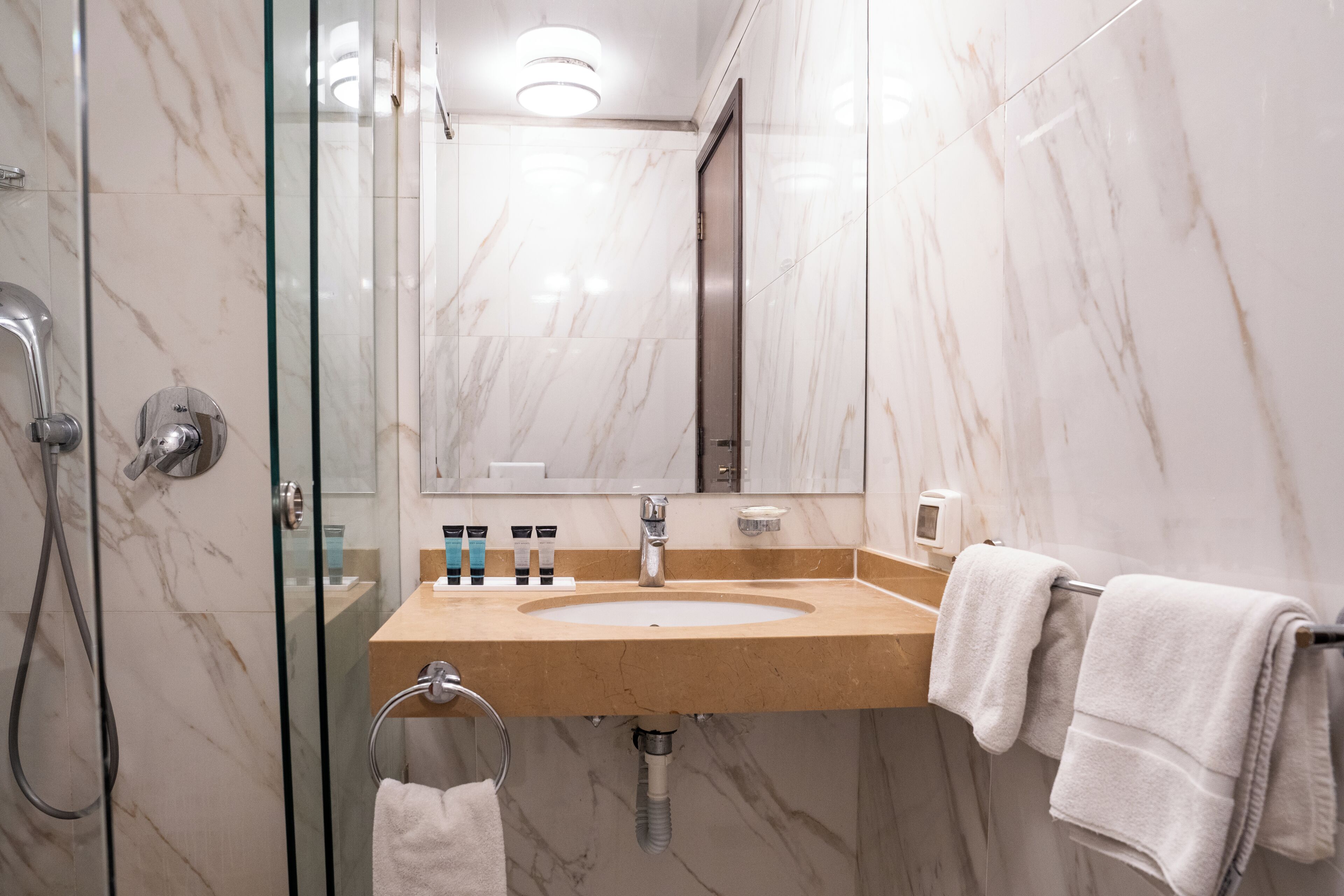 Un baño con ducha de vidrio, pared con azulejos de mármol, lavamanos con toallero y espejo.