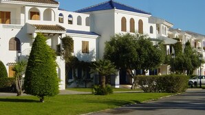A row of white buildings with blue roofs and arched windows, surrounded by well-maintained greenery and a clear sky. - Apartamentos Mar Menor (San Javier)