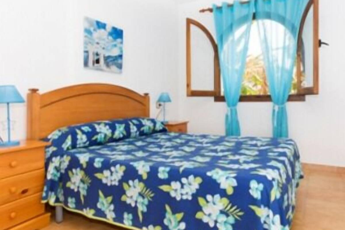 Une chambre à coucher avec un lit en bois, une couvre-lit à motifs floraux bleus, une table de chevet avec une lampe et une fenêtre avec des rideaux bleus.