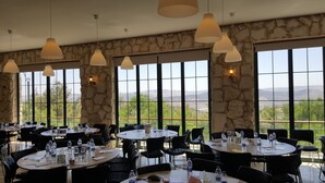 Un restaurante con mesas y sillas redondas, amplias ventanas con vistas a la montaña, paredes de piedra y lámparas colgantes.