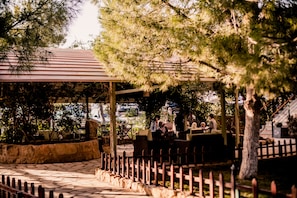 Un área de estar al aire libre con mesas y sillas bajo una pérgola cubierta, rodeada de árboles y un muro de piedra.