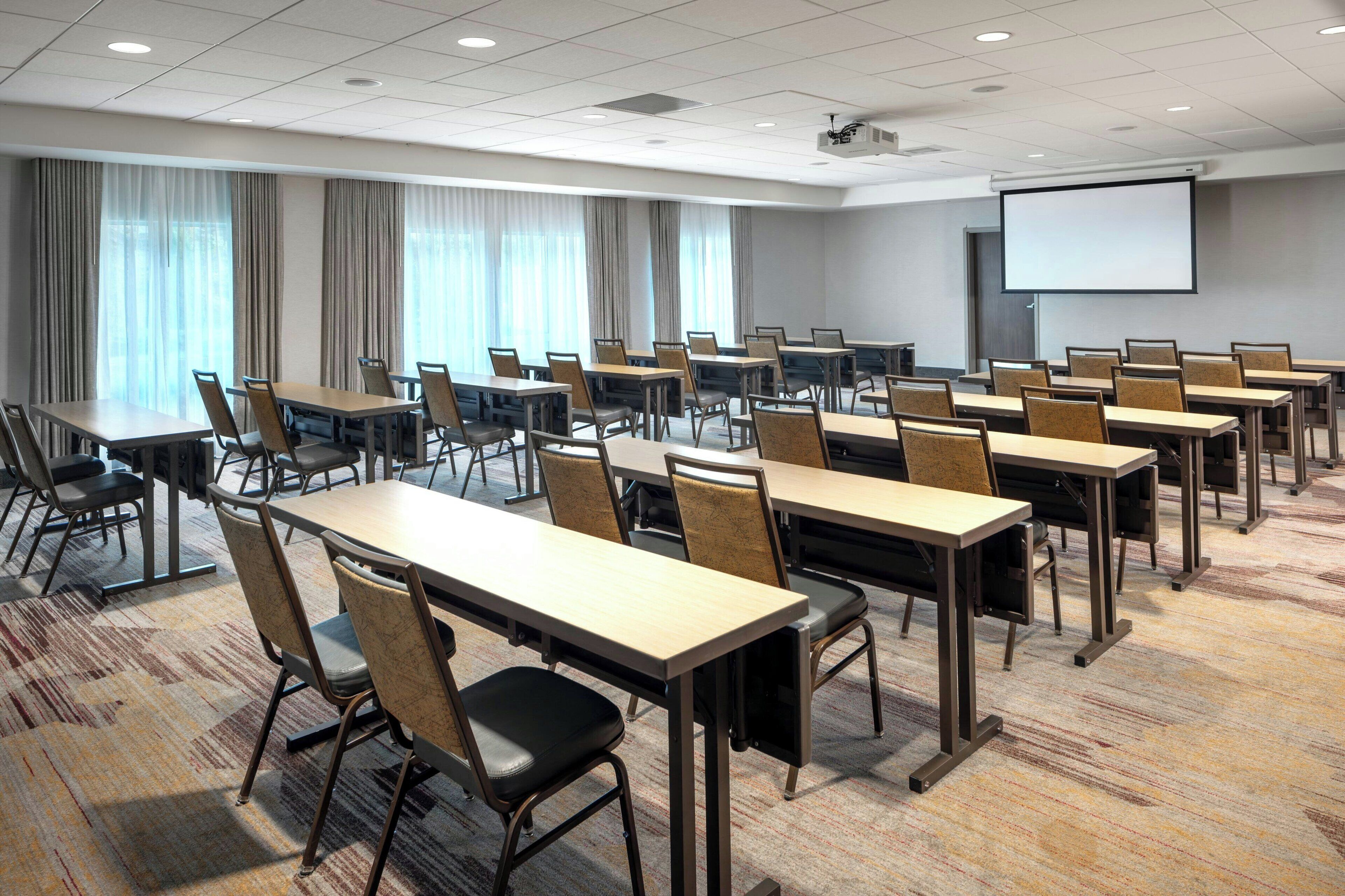 A conference room with multiple tables and chairs, a projector screen, and large windows with curtains.