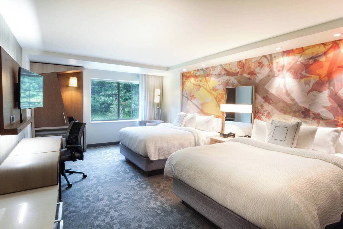 A hotel room with two beds, a desk, a chair, a TV, and a large window with a view of trees.
