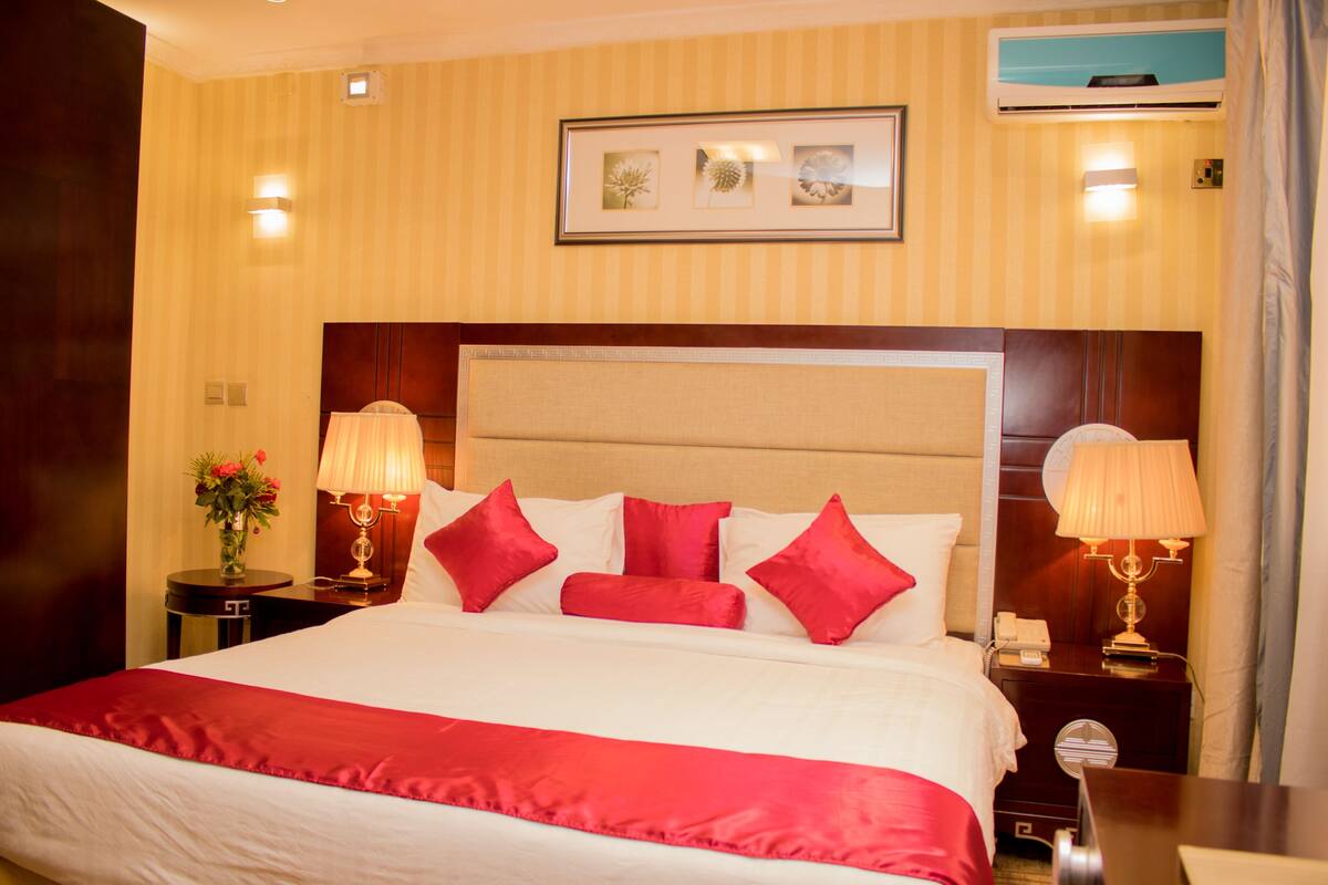 A hotel room with a large bed, red and white bedding, two bedside tables with lamps, a framed picture on the wall, and an air conditioning unit.