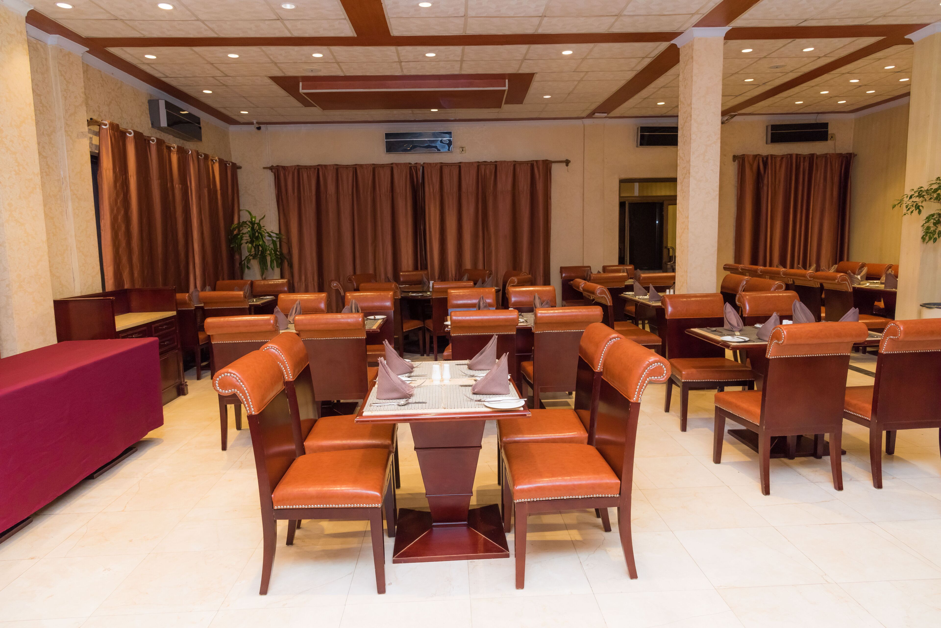 A spacious dining area with multiple tables and chairs, a red counter, and brown curtains.