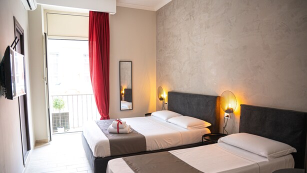 A hotel room with two beds, a balcony, a red curtain, a wall-mounted TV, and a bedside lamp. - Napoli City Rooms (Naples)