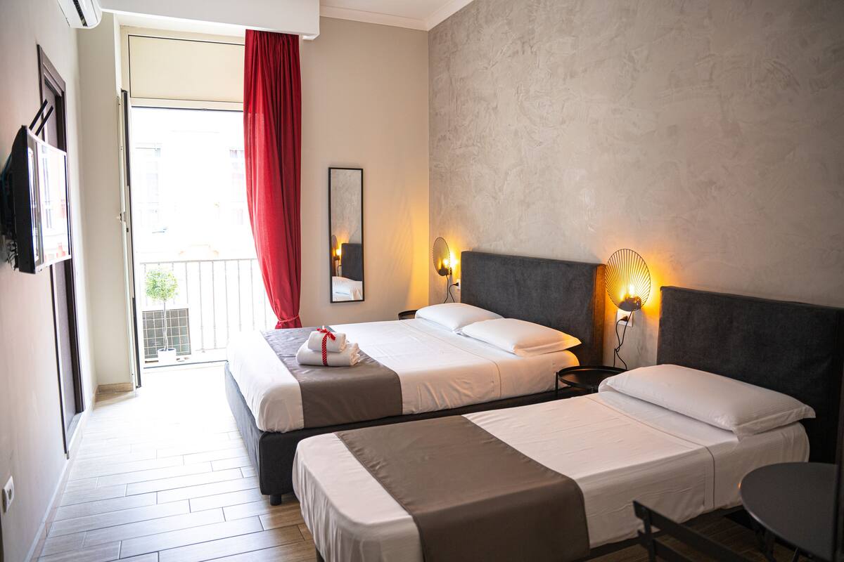 Habitación de hotel con dos camas, un balcón, una cortina roja, un televisor montado en la pared y una lámpara de noche.