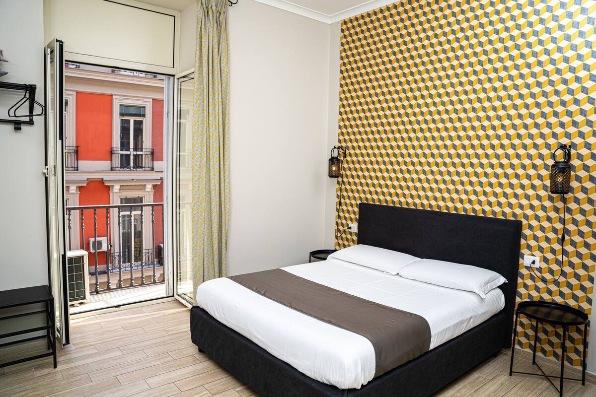 Un dormitorio con cama, mesita de noche, lámpara de pared y vistas a un balcón exterior.