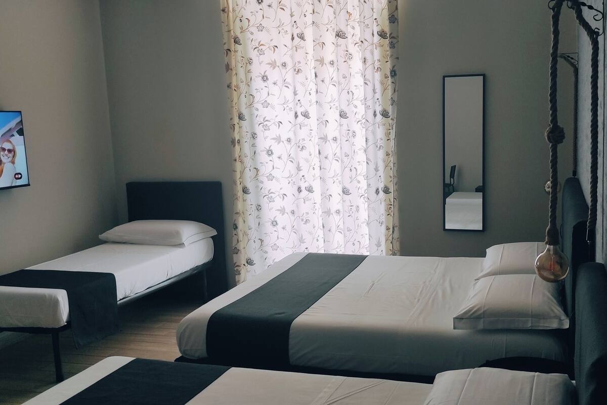 Habitación de hotel con dos camas individuales, un televisor en la pared, un espejo y una ventana con cortinas de flores.