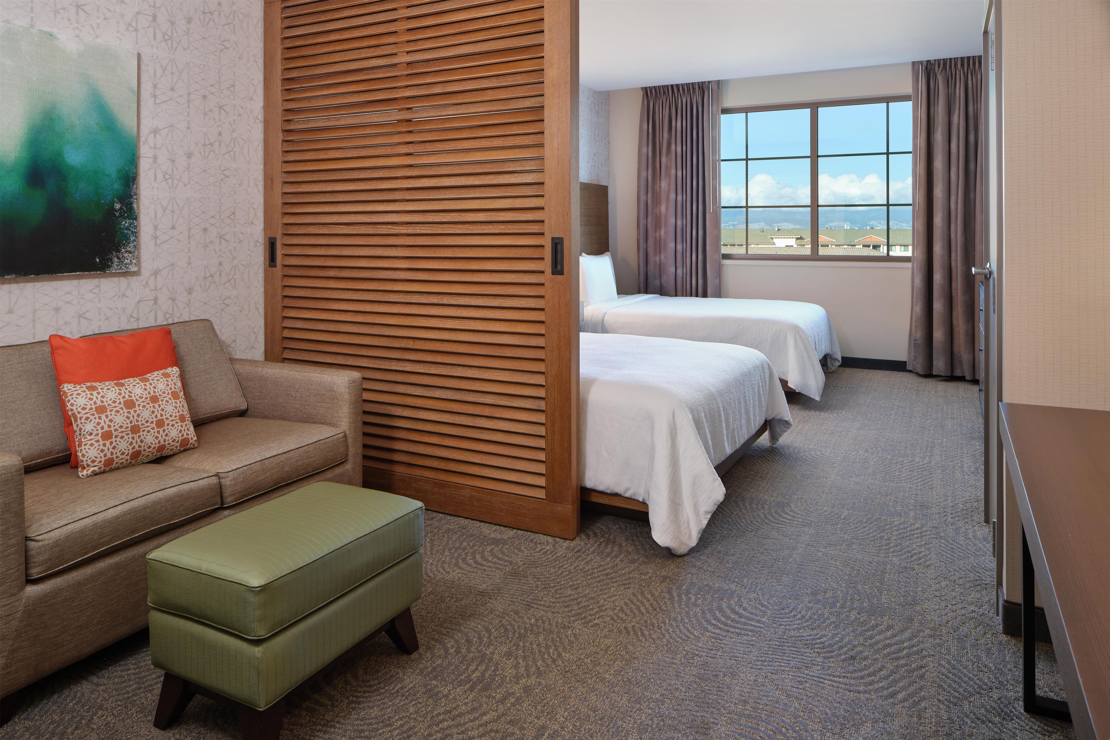 A hotel room with a sofa, two beds, a wooden partition, and a view of the sky.
