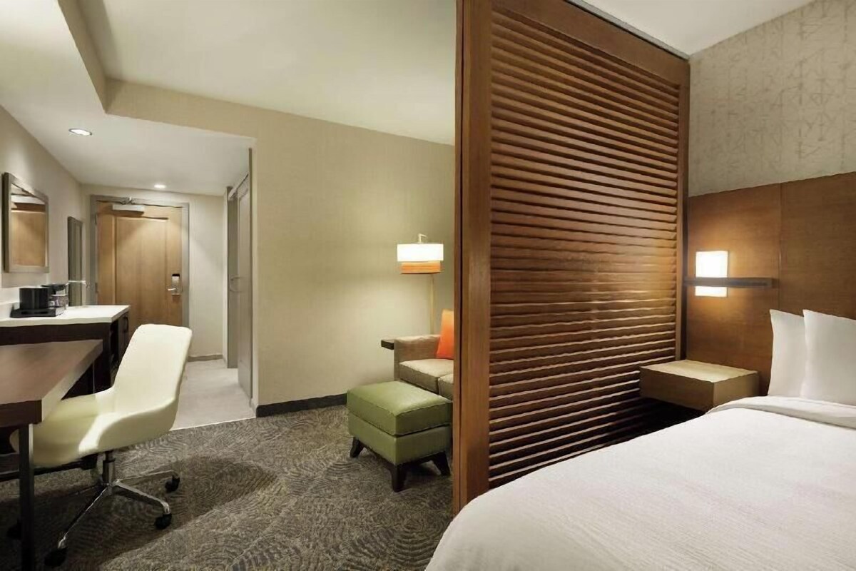 Hotelkamer met een bed, een bureau met stoel, een kleine tafel met lamp en een scheidingswand met horizontale latten.