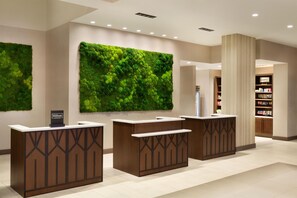 A hotel reception area with three reception desks, a large green wall, and a bookshelf with magazines.
