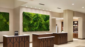 A hotel reception area with three wooden reception desks, a large green moss wall, and a bookshelf in the background. - Embassy Suites By Hilton Oahu Kapolei (Kapolei)
