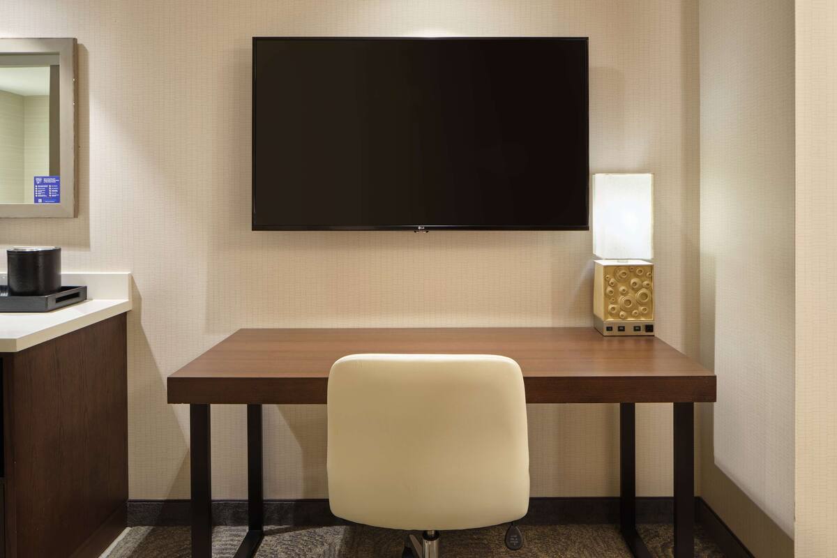Een hotelkamer met een flatscreen televisie aan de muur, een houten bureau, een witte stoel, een lamp en een minibar.