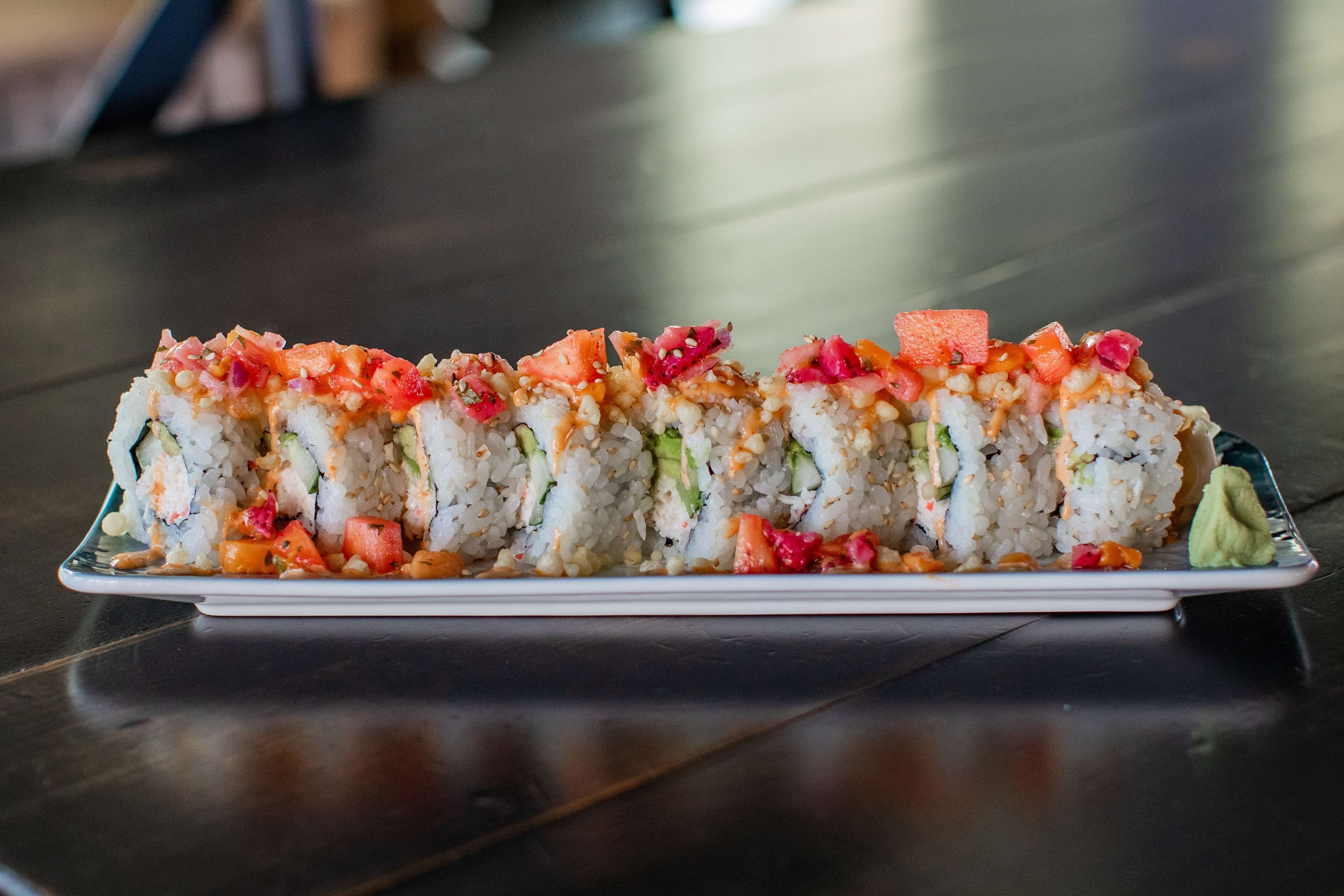 A plate of sushi rolls with various toppings and a small dollop of wasabi on a dark wooden table.