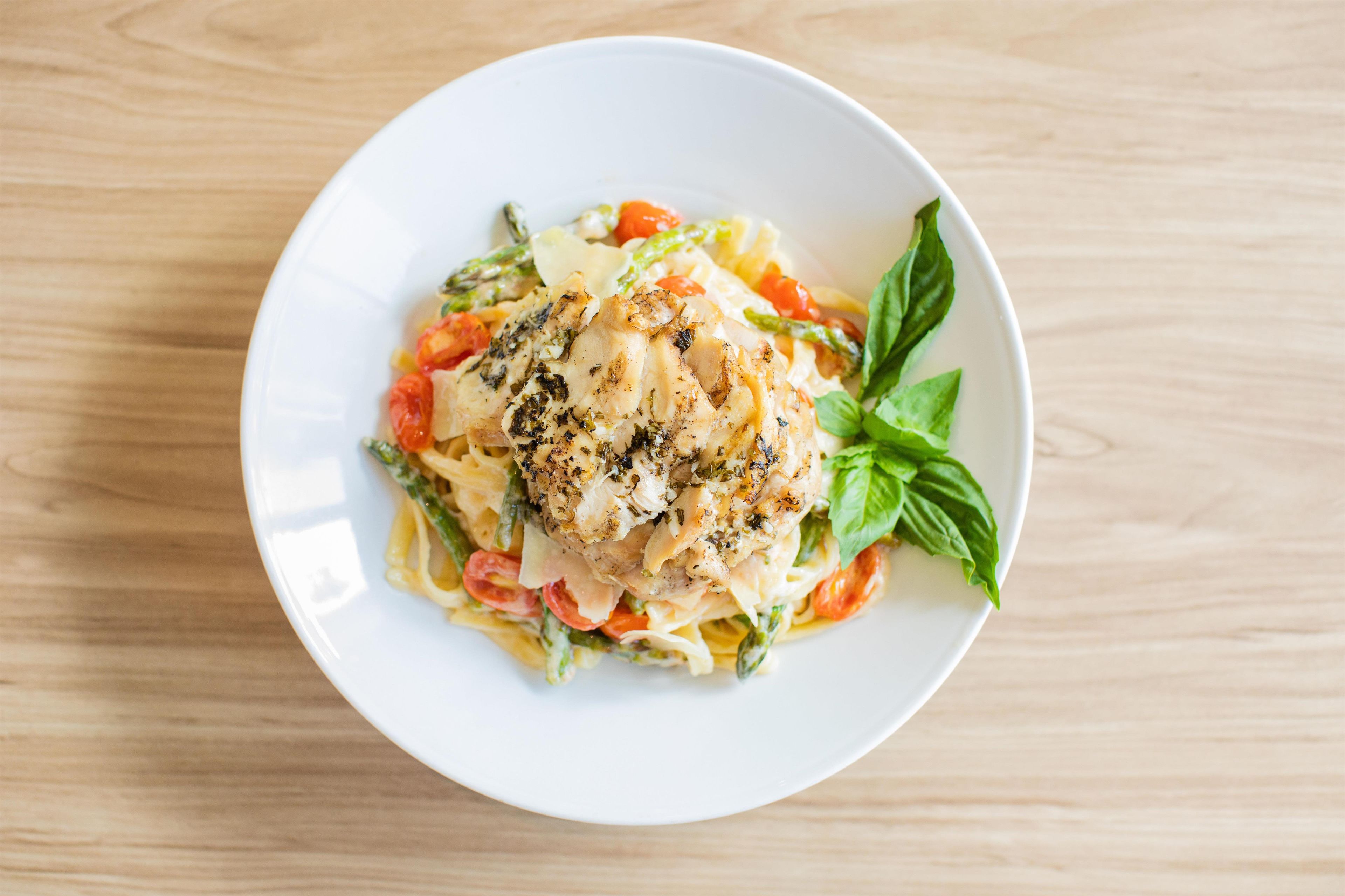 Un plato blanco con pasta, pollo y verduras sobre una mesa de madera.