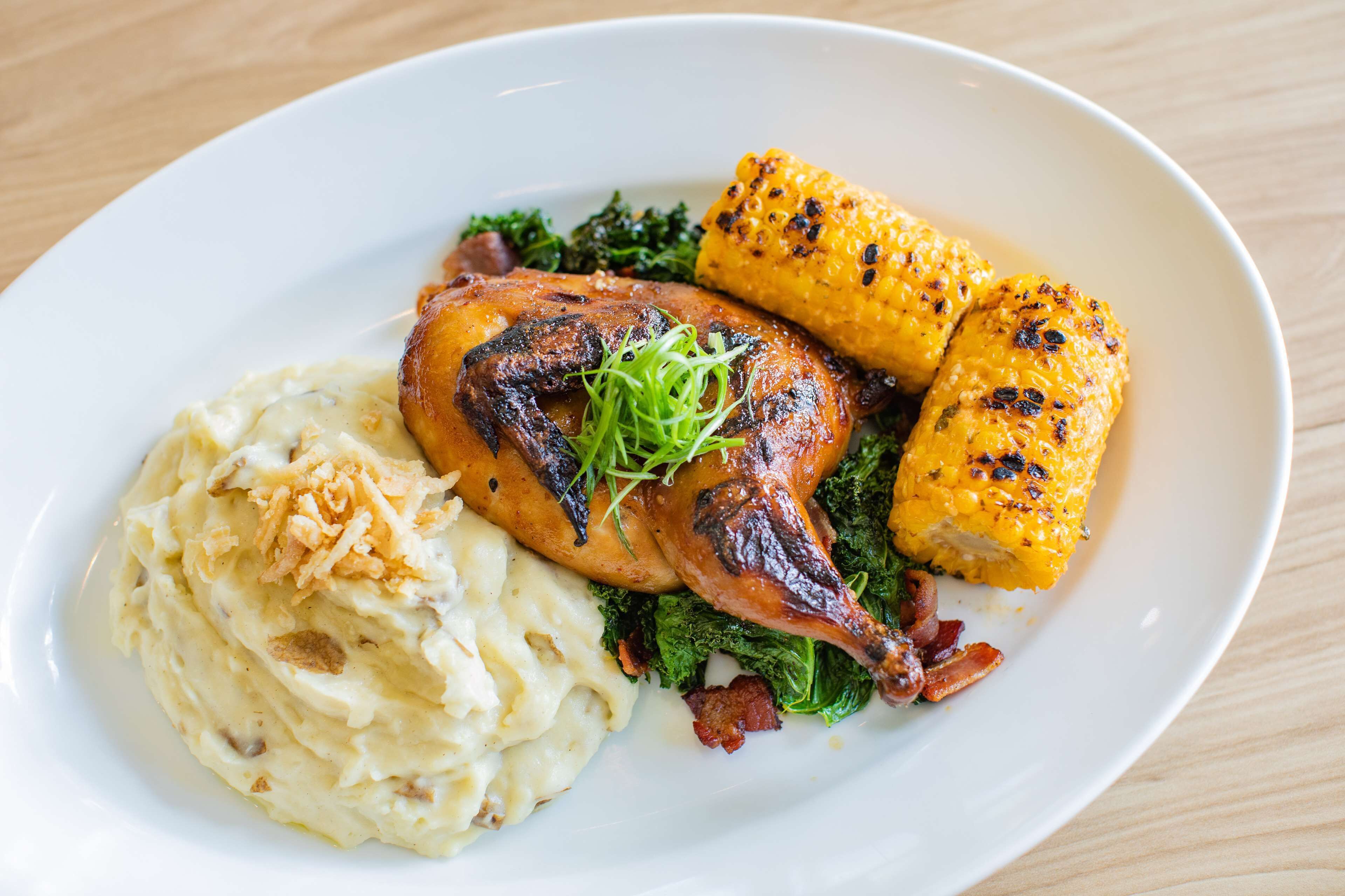 A plate with roasted chicken, mashed potatoes, kale, and grilled corn on the cob.