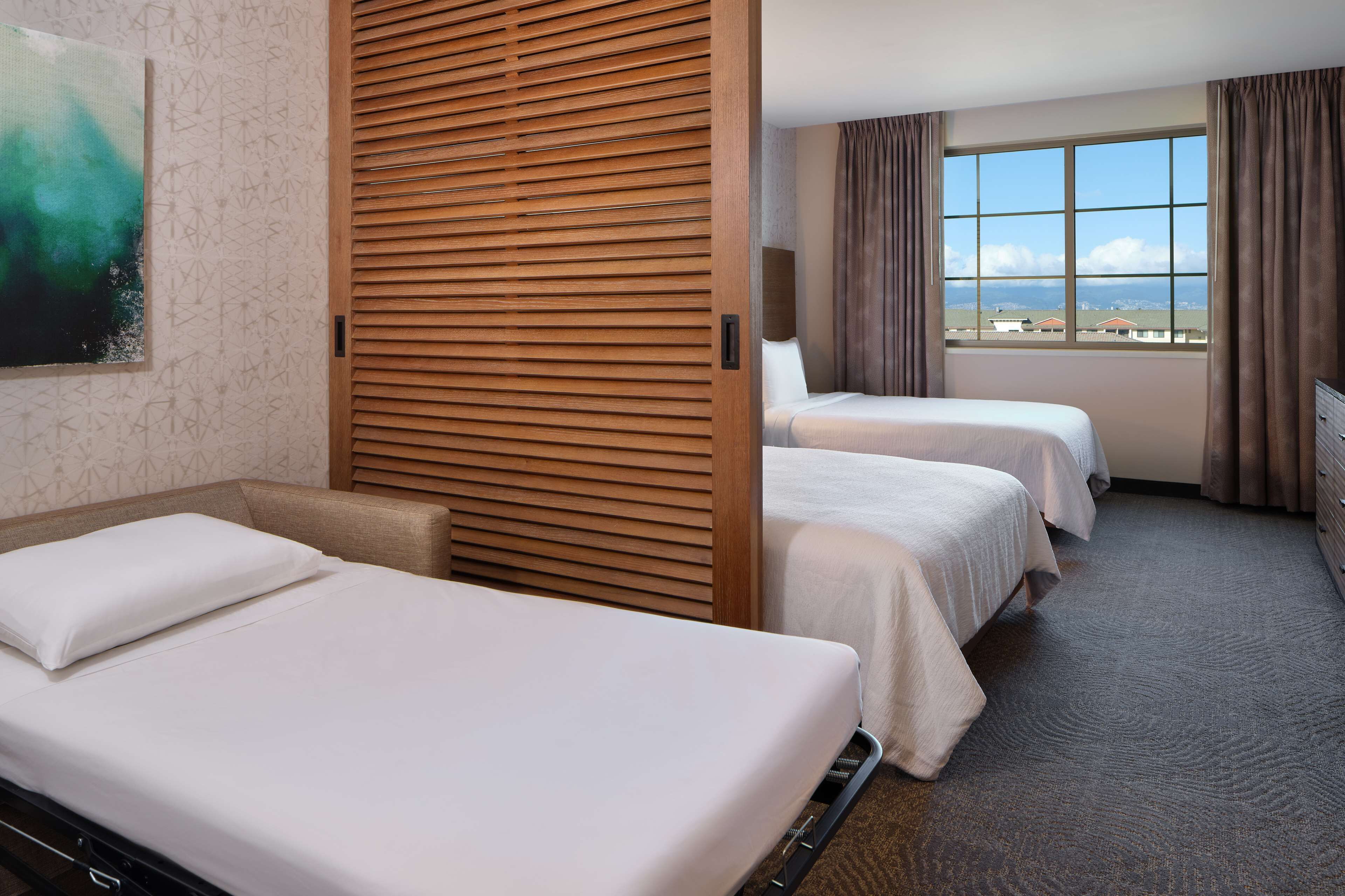 Habitación de hotel con dos camas, un separador de madera, un ventanal con vistas y un cuadro en la pared.