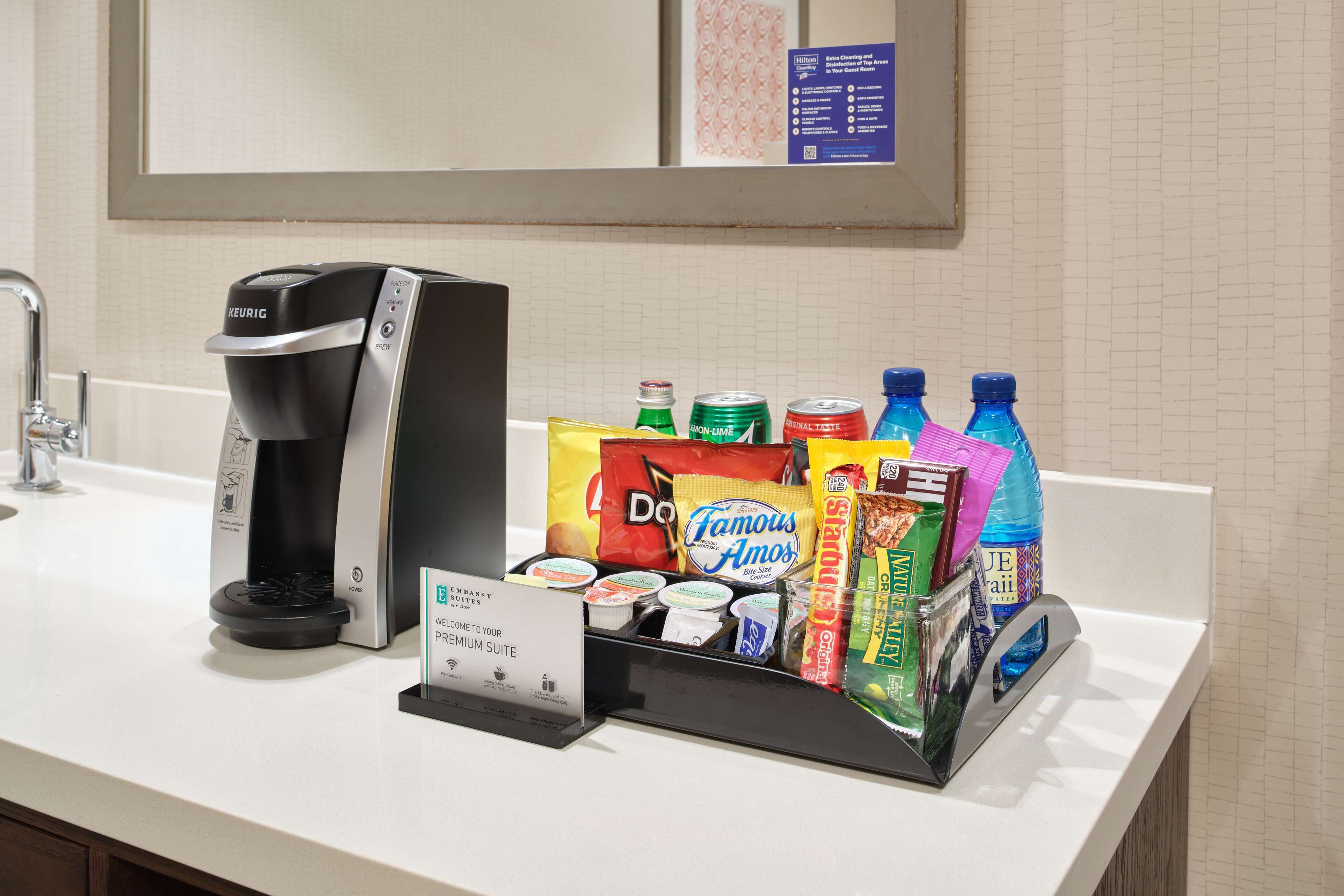A hotel coffee station with a Keurig coffee maker, a variety of beverages including bottled water, soda, and juice, and a sign indicating a premium suite.
