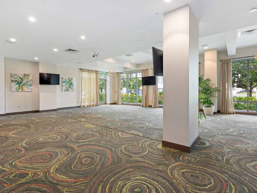 A spacious hotel room with a patterned carpet, large windows, and a flat-screen TV mounted on the wall.