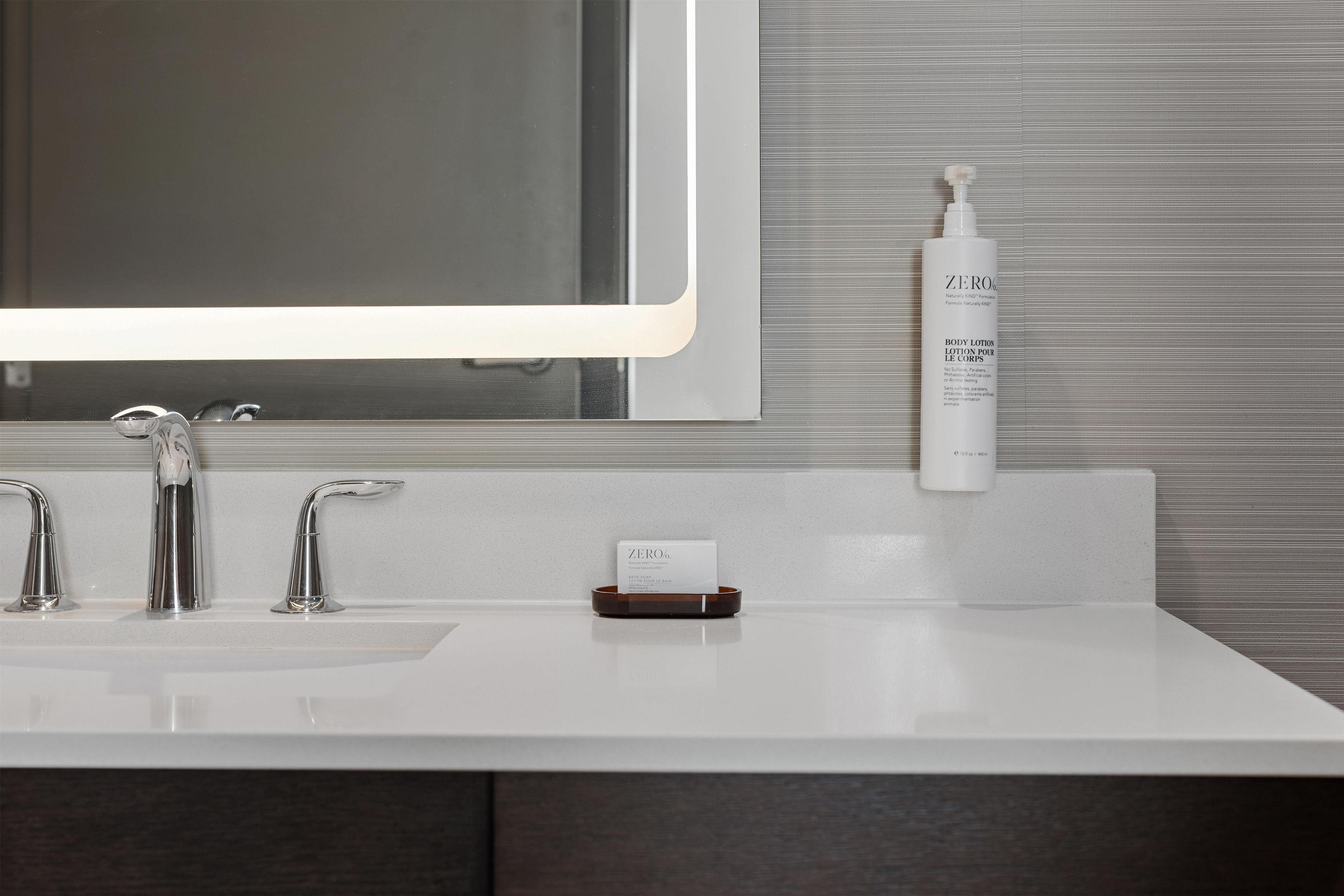 Un baño con un lavabo blanco, dos grifos, un espejo y una botella de crema corporal.