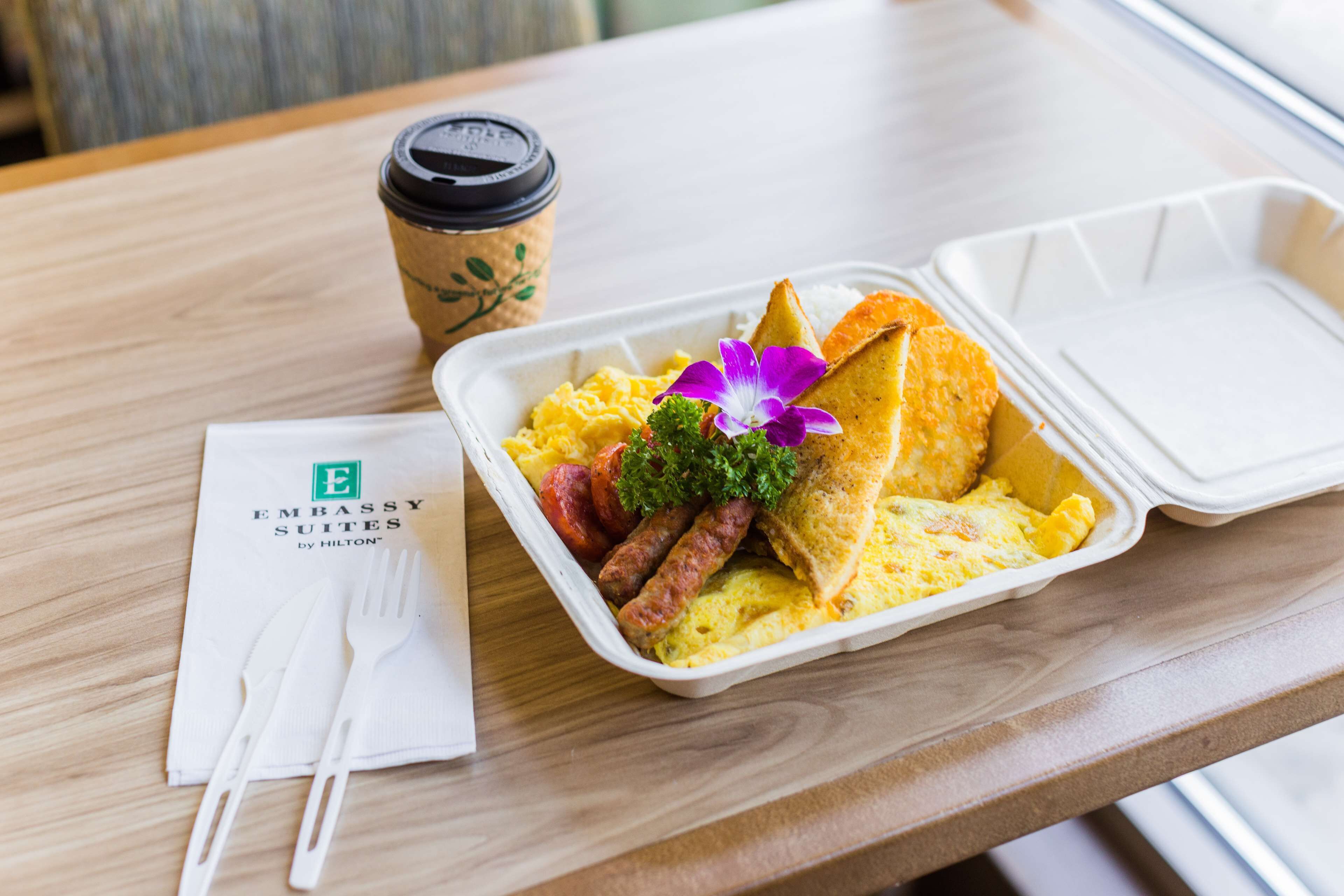 A meal in a takeout container with scrambled eggs, sausages, toast, and a purple flower garnish, accompanied by a coffee cup and a napkin with a logo.