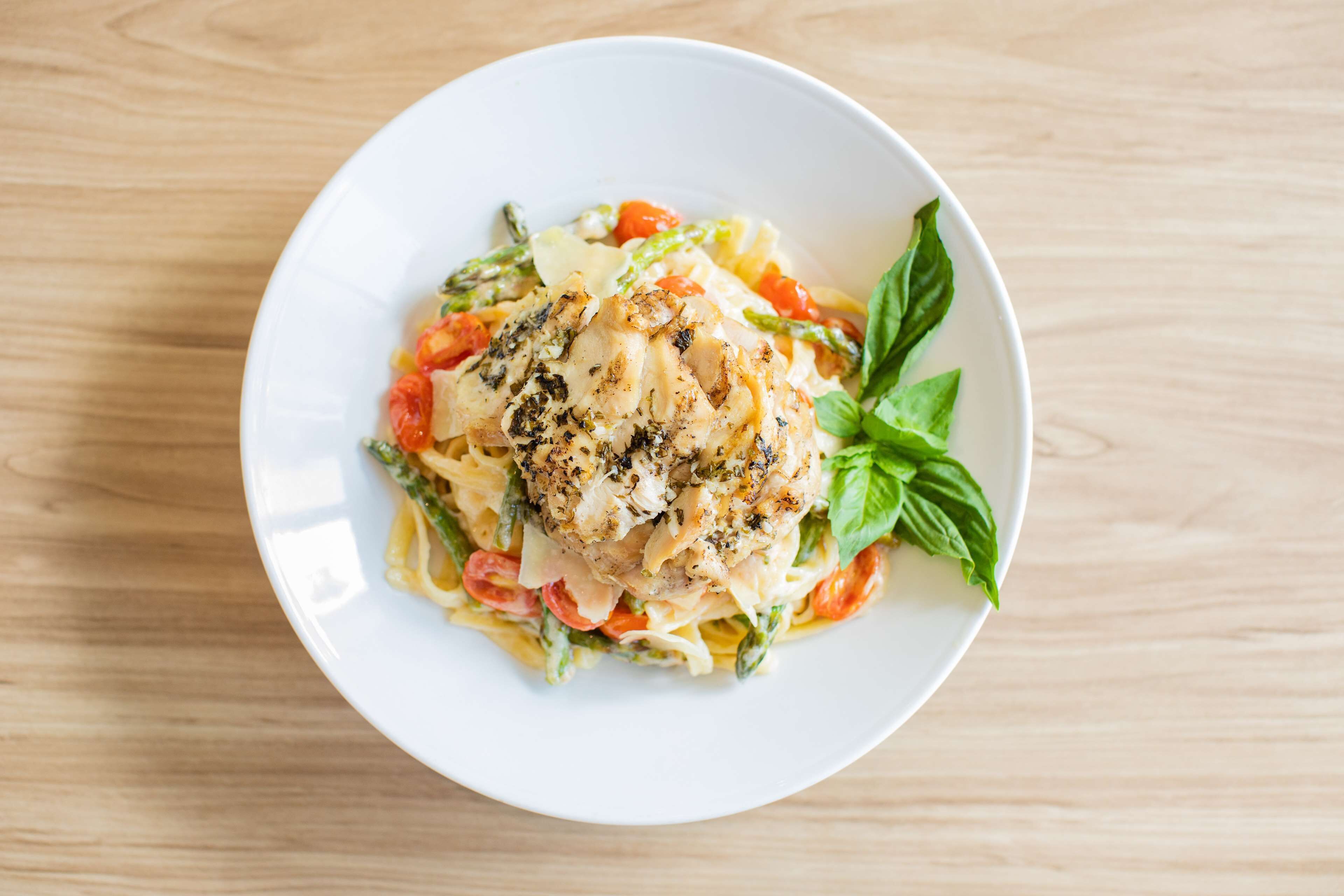 Un plato blanco con pasta, pollo, espárragos y tomates sobre una mesa de madera.