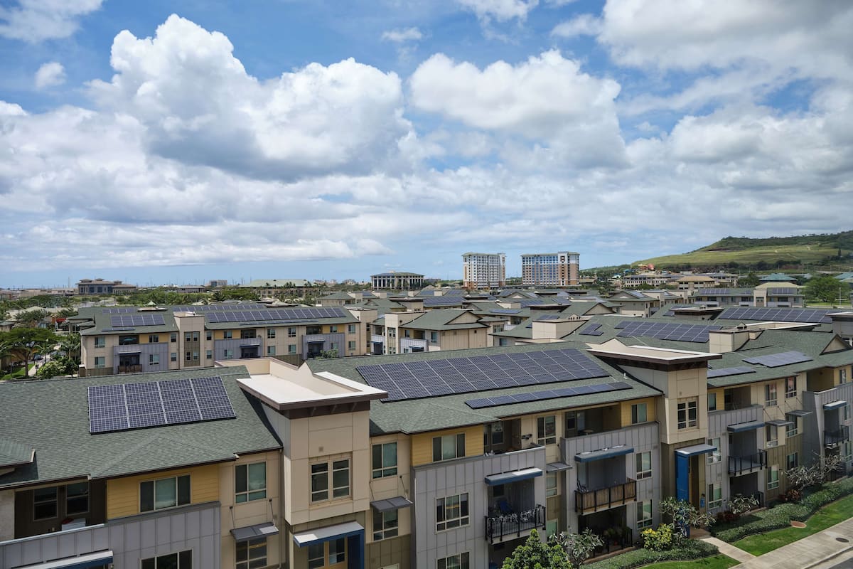 Een woongebied met moderne appartementencomplexen, zonnepanelen op de daken en een heuvelachtig landschap op de achtergrond.