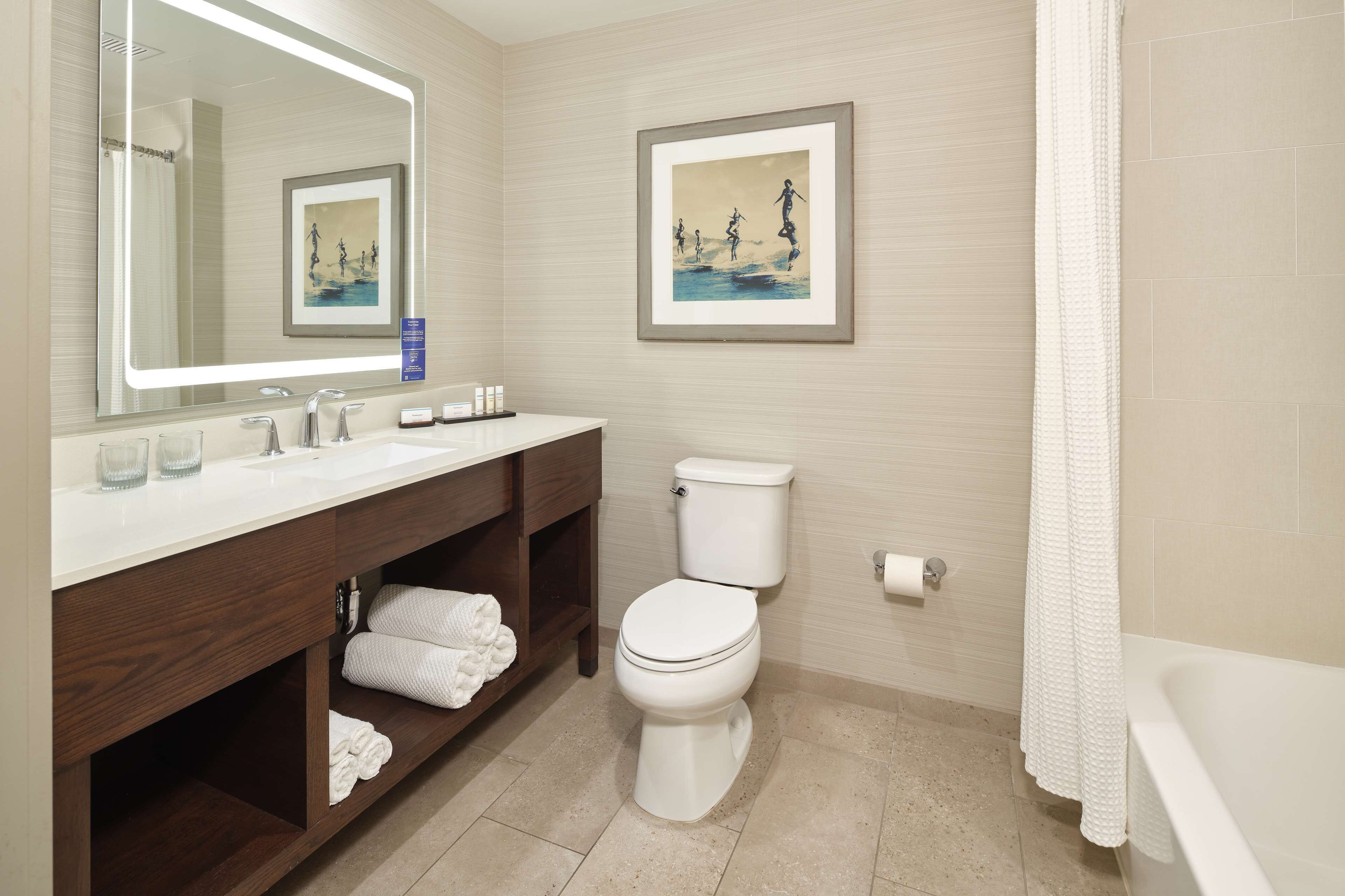 A modern bathroom with a white sink, vanity, and toilet. There are two framed artworks on the wall, a towel rack, and a white bathtub.