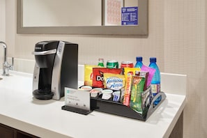 Een hotelminibar met een Keurig koffiezetapparaat, diverse drankjes, snacks en een bordje dat een ambassadesuite aangeeft.
