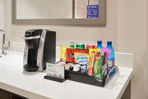 Un minibar de hotel con una cafetera Keurig, diversas bebidas, aperitivos y un letrero que indica una suite embajada.