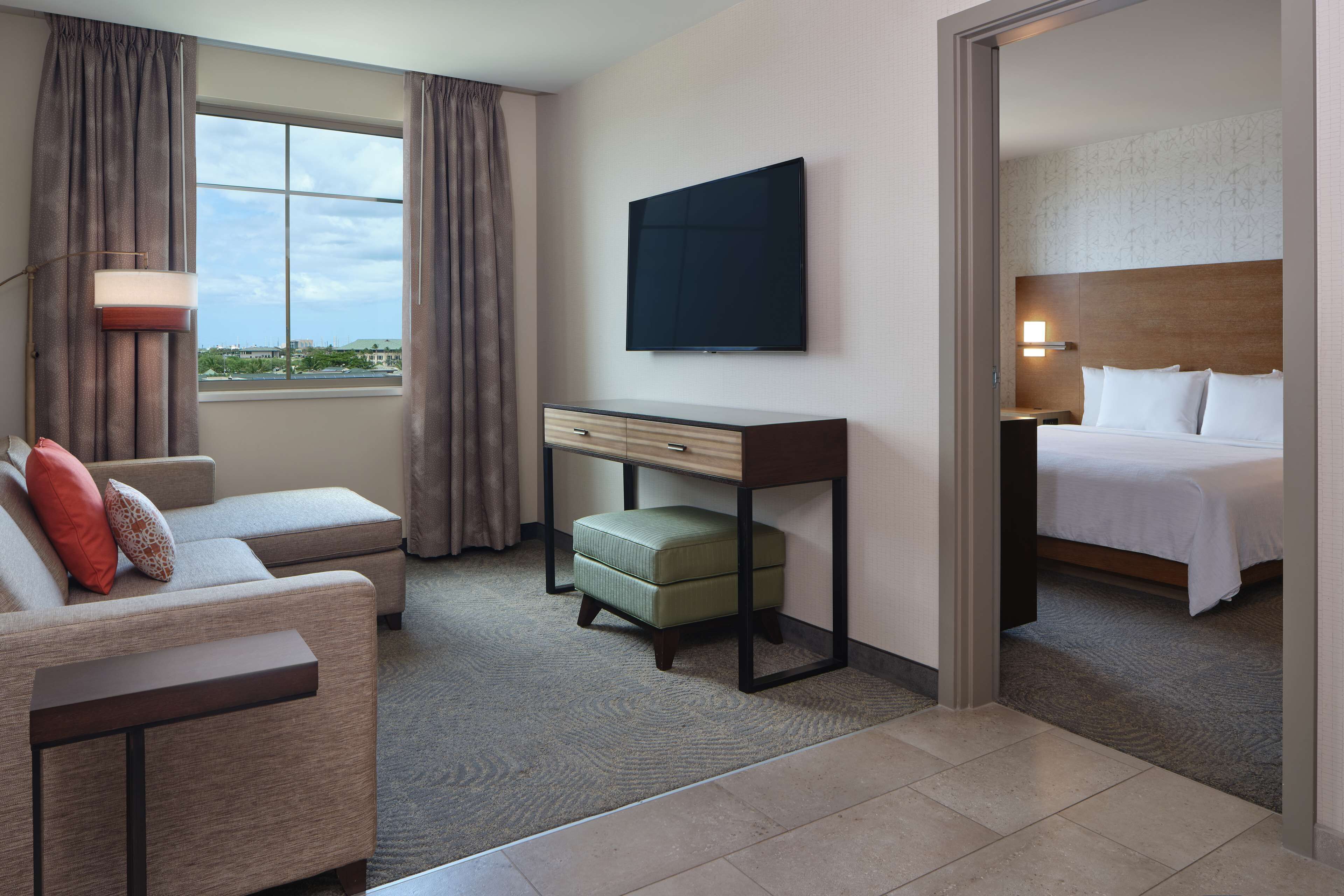 A hotel room with a bed, a desk, a chair, a small ottoman, a TV, and a window with curtains.