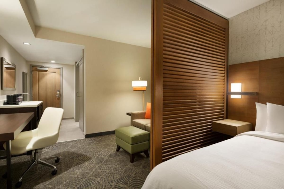 Hotelkamer met een bed, een bureau met stoel, een klein tafeltje en een wandlamp.