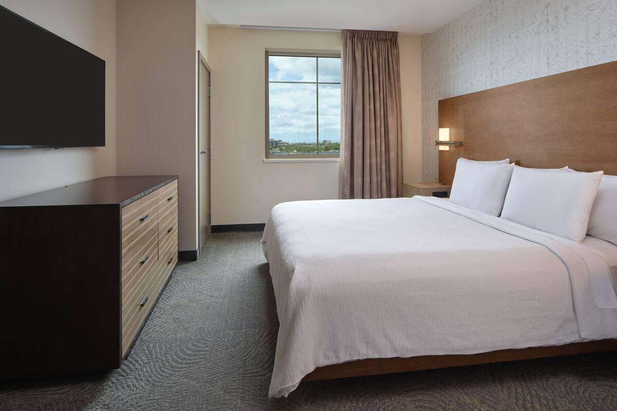 Een hotelkamer met een bed, een dressoir, een televisie, een raam met gordijnen en een nachtkastje met een lamp.