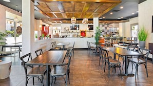 A modern restaurant interior with wooden tables and chairs, a kitchen area, and potted plants. - Embassy Suites By Hilton Oahu Kapolei (Kapolei)