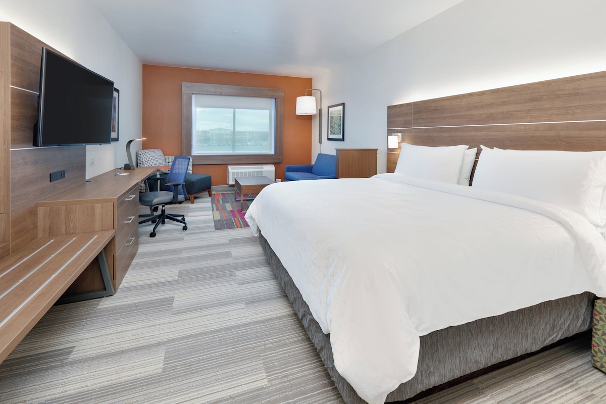 A hotel room with a large bed, a desk with a flat-screen TV, a chair, a window, and a headboard.