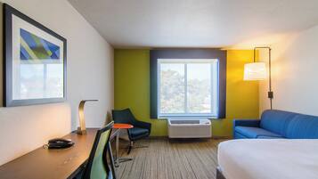 A hotel room with a bed, a desk with a chair, a window with a view, and a wall-mounted lamp.