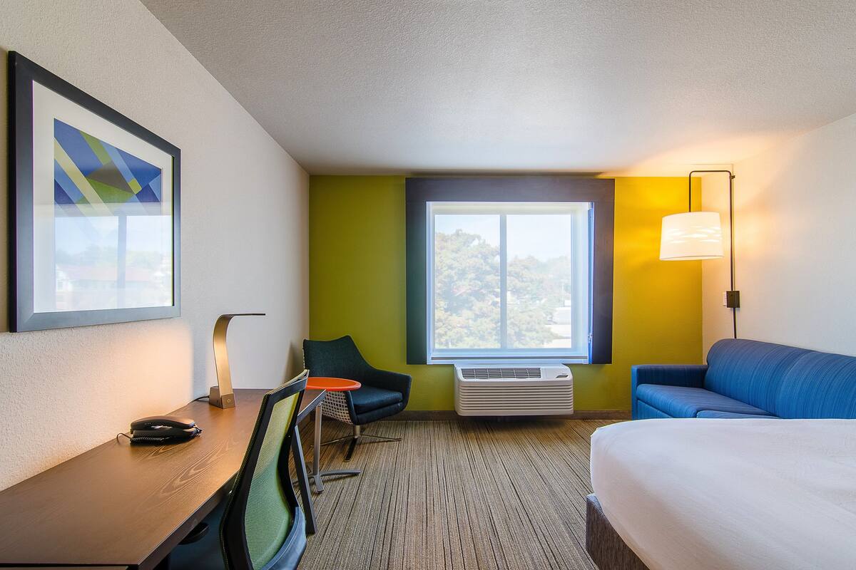A hotel room with a bed, a desk with a chair, a small table, a lamp, and a large window with a view.