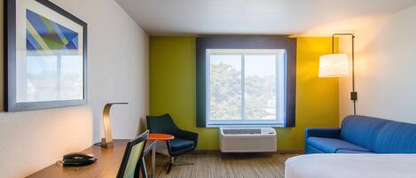 Habitación de hotel con cama, escritorio con silla, mesita, lámpara y un ventanal con vistas.