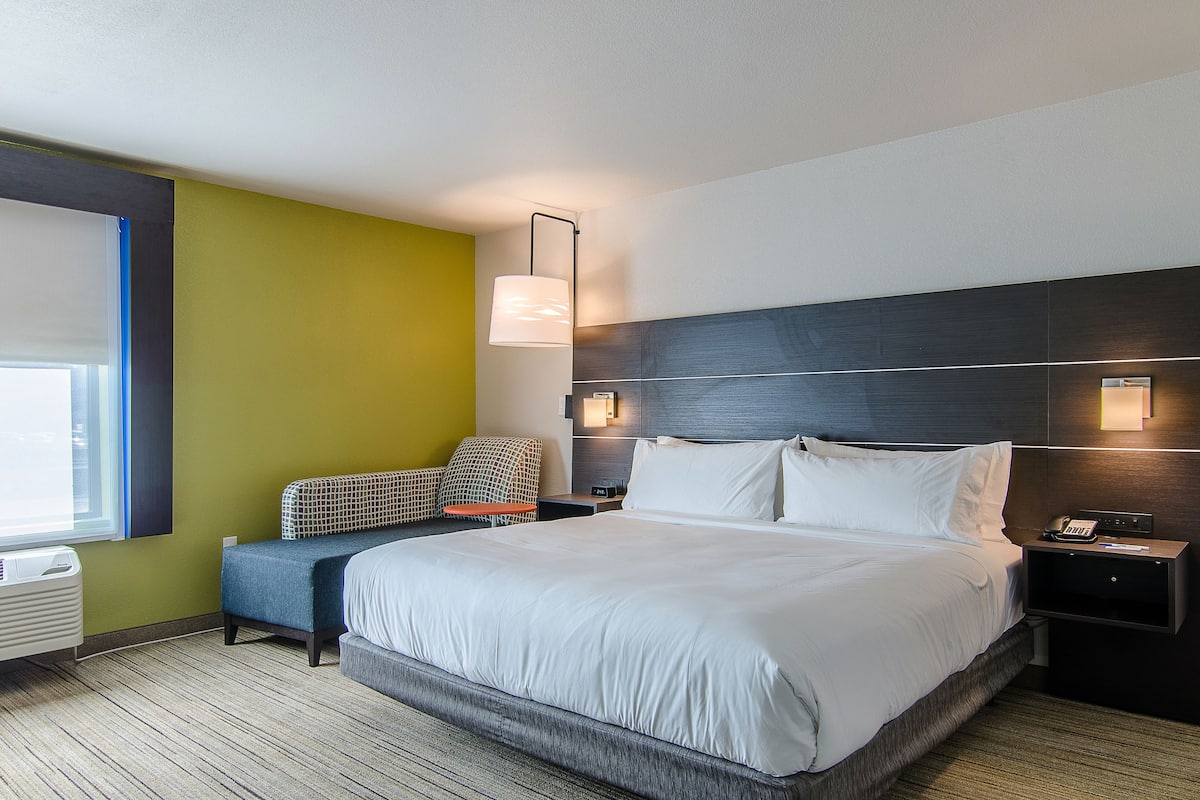 A modern hotel room with a large bed, a bedside table with a lamp, a chair, and a window with blinds.