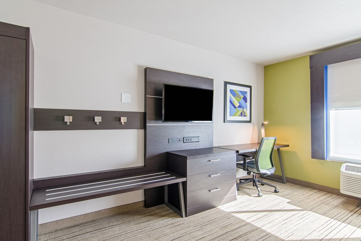 A modern hotel room with a dark wood desk, a flat-screen TV, a green chair, and a window with blinds.