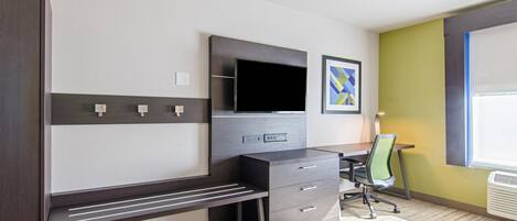 A modern hotel room with a dark wood desk, a flat-screen TV, a green chair, and a window with blinds.