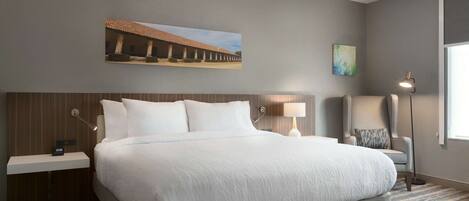 Habitación de hotel con una cama grande, una mesita de noche, una silla, una lámpara de pie y un cuadro en la pared.