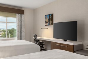 Ein Hotelzimmer mit zwei Betten, einem Schreibtisch, einem Stuhl, einem Flachbildfernseher und einem Bild an der Wand.