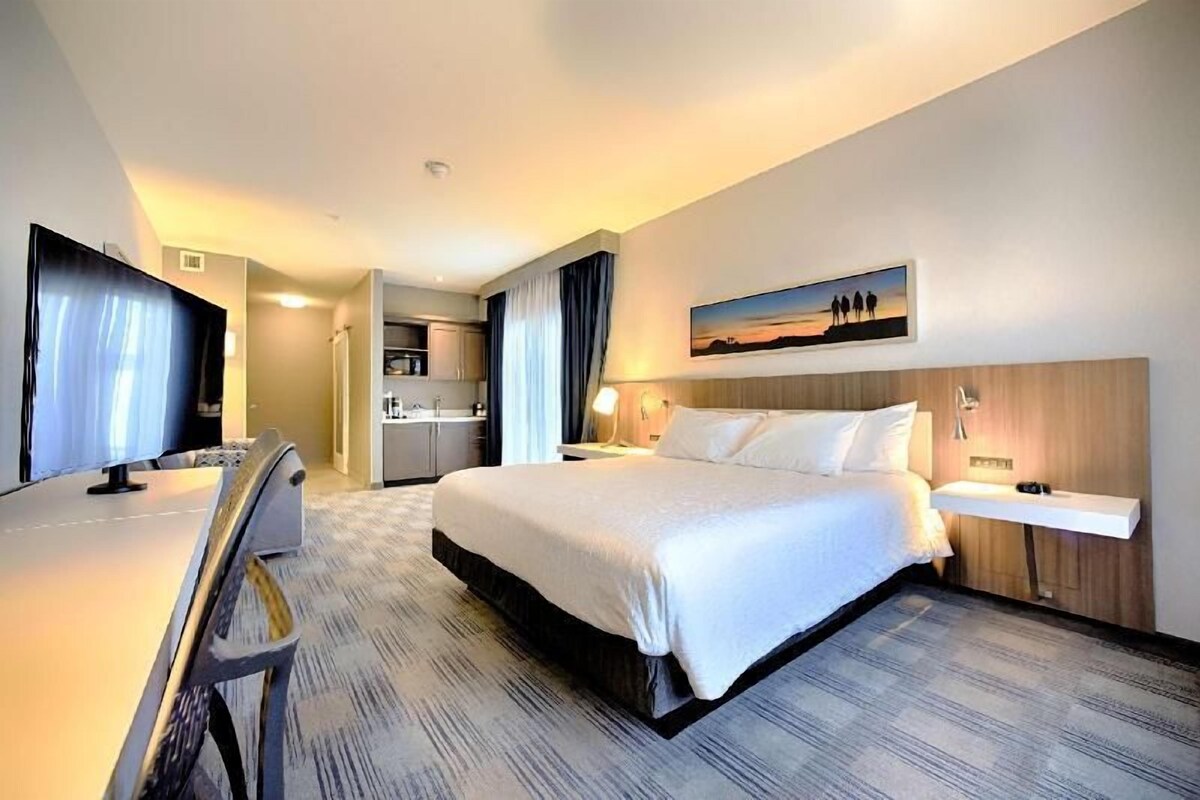 A hotel room with a bed, a desk, a TV, a painting, and a kitchenette.