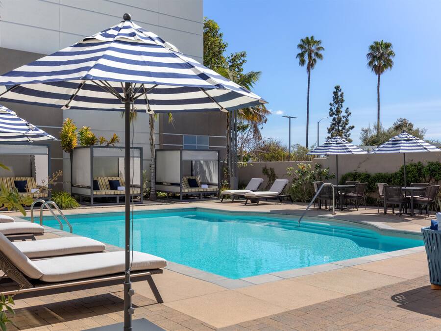 A pool area with lounge chairs and umbrellas, surrounded by palm trees and a modern building.
