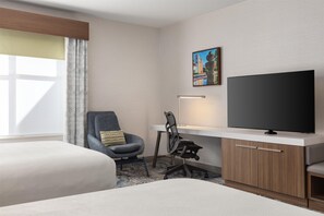 Ein Hotelzimmer mit zwei Betten, einem Schreibtisch, einem Flachbildfernseher, einem Sessel und einem Bild an der Wand.