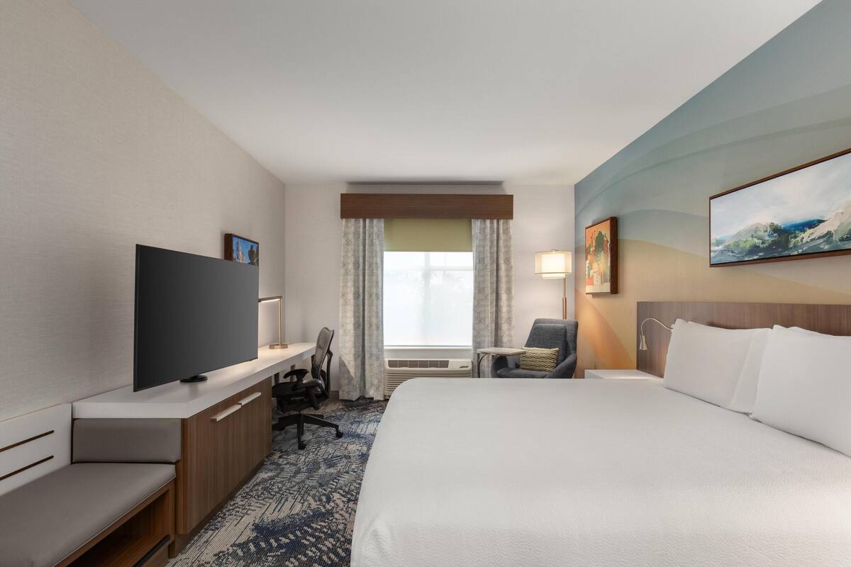 Une chambre d’hôtel comprenant un lit, un bureau avec un téléviseur à écran plat, une chaise, une petite table et une fenêtre avec des rideaux.