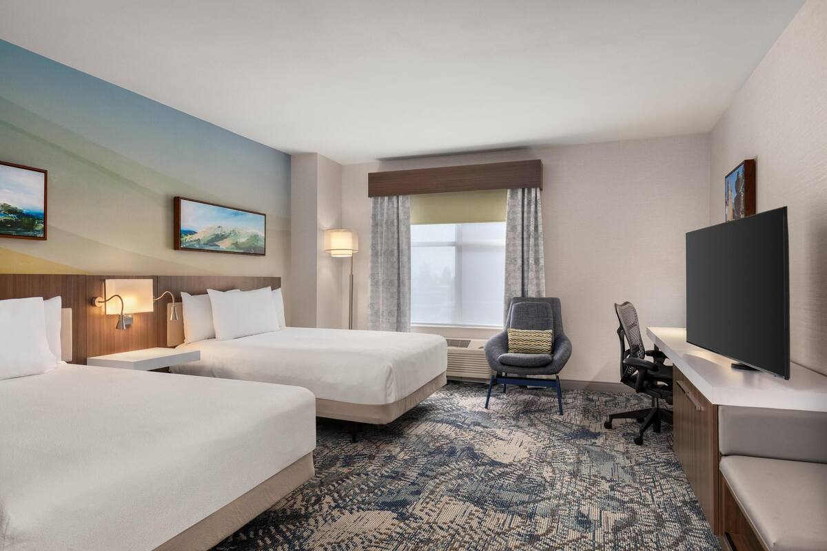 Une chambre d’hôtel avec deux lits, un bureau doté d’un téléviseur à écran plat, une chaise, une lampe et une fenêtre avec des rideaux.